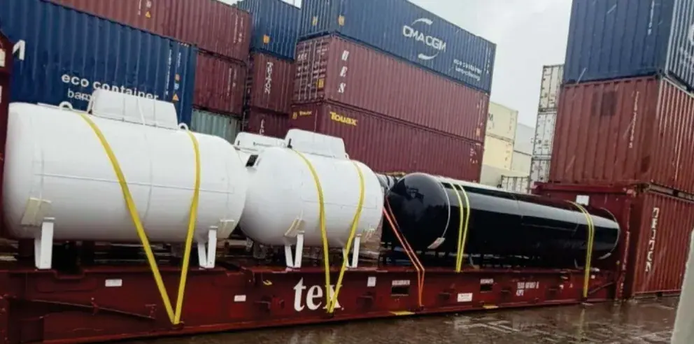Three tanks strapped down on a red flat-rack in front of stacked shipping containers