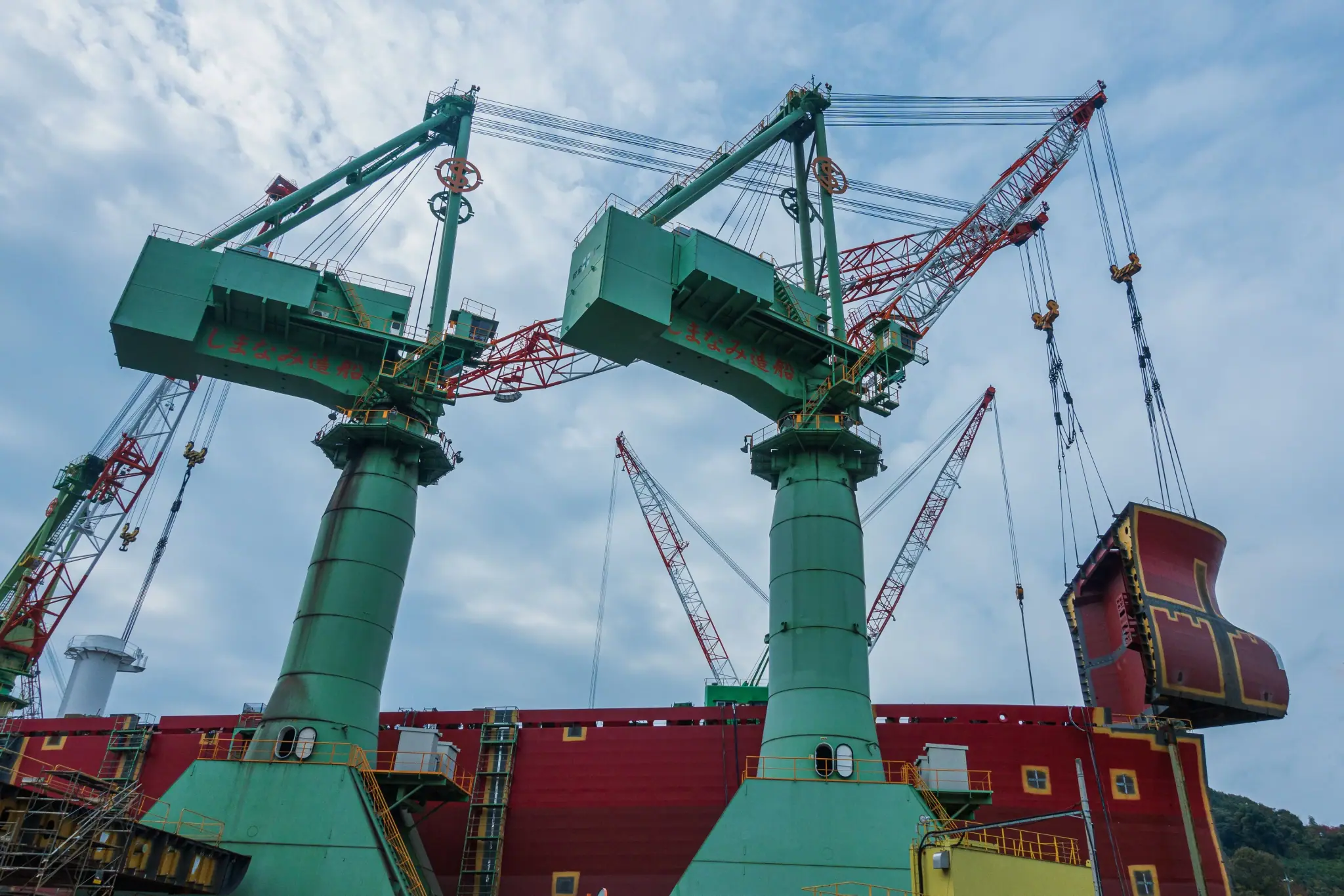 Two large green cranes lifting a huge steel block at a shipyard
